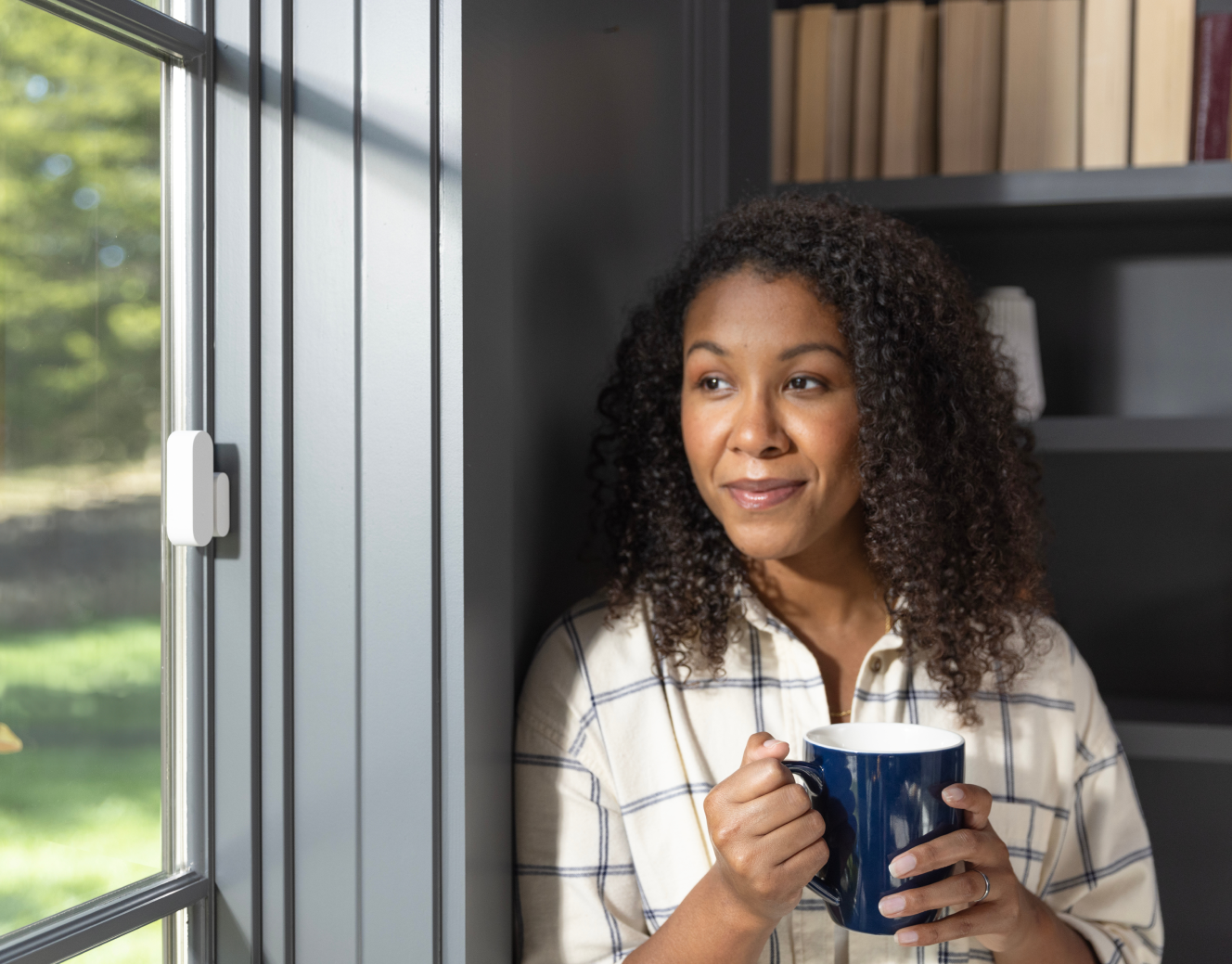 Woman drinking out of a mug and staring out the window with an ADT door/window sensor on it