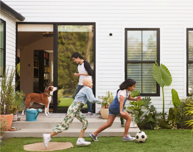 Family playing in the yard with a Google Nest Cam (outdoor or indoor, battery) installed on their home